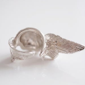 May include: A silver-colored ring with a wing design. The ring features detailed feather textures on the wings, creating a realistic appearance. The ring is set against a white background.