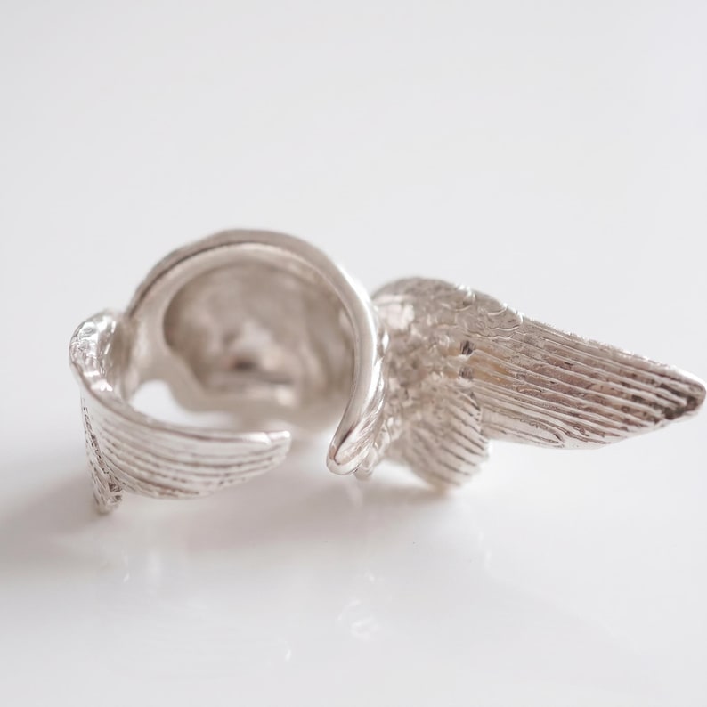 May include: A silver-colored ring with a wing design. The ring features detailed feather textures on the wings, creating a realistic appearance. The ring is set against a white background.