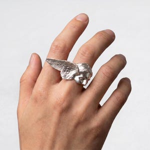 May include: A silver ring featuring a detailed angel's head with feathered wings. The ring is worn on a finger, showcasing the intricate design and metallic sheen. The angel's face is sculpted with delicate features.