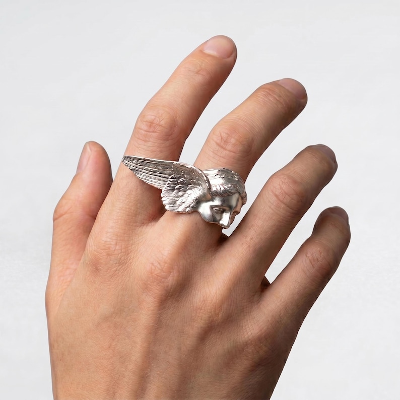 May include: A silver ring featuring a detailed angel's head with feathered wings. The ring is worn on a finger, showcasing the intricate design and metallic sheen. The angel's face is sculpted with delicate features.