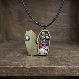 Peut inclure: Pendentif en forme de cercueil en laiton antique. Le cercueil ouvert révèle une collection de pierres précieuses colorées, de perles et une petite couronne. Le collier est suspendu à un cordon noir. Un bijou unique.