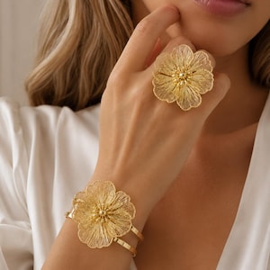 May include: A gold-toned metal ring and a matching cuff bracelet, both featuring a large, intricate flower design with delicate, wire-like filigree petals and a textured, beaded center. The jewelry set displays a bright, polished finish.
