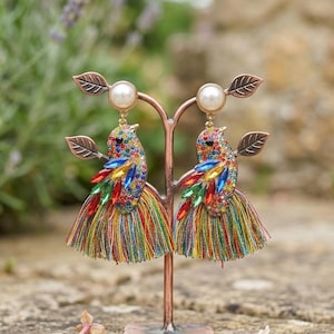 May include: A pair of colorful bird-shaped earrings. Each earring features a pearl at the top, a body adorned with multi-colored rhinestones, and a tassel skirt. The earrings are displayed on a copper-colored stand with leaf accents.