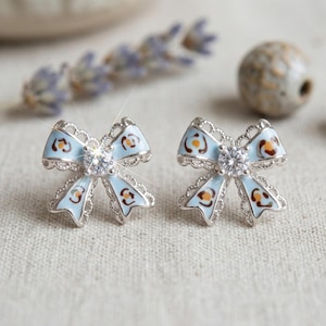 May include: A pair of bow-shaped stud earrings. The earrings feature a light blue enamel design with brown accents and a clear, round gemstone in the center. The edges are detailed with a silver filigree.