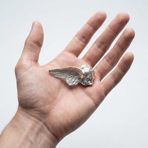May include: A silver-colored winged head sculpture, possibly a brooch or pendant, is displayed. The sculpture features detailed wings and a classical face. The item is held in a hand against a white background, showcasing the intricate design and craftsmanship.