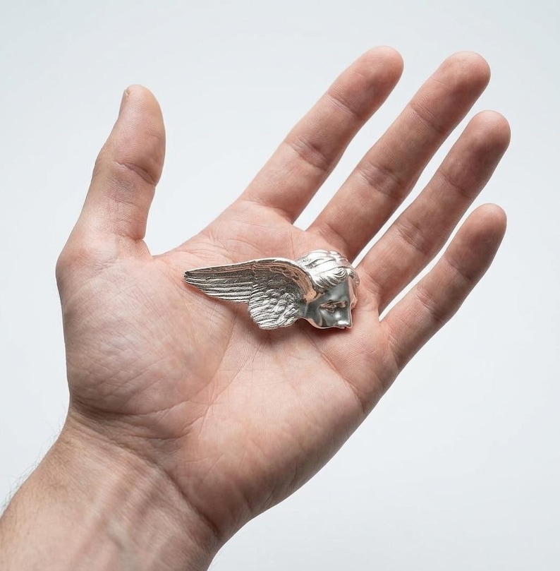 May include: A silver-colored winged head sculpture, possibly a brooch or pendant, is displayed. The sculpture features detailed wings and a classical face. The item is held in a hand against a white background, showcasing the intricate design and craftsmanship.