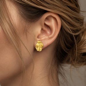 May include: A gold-toned stud earring shaped like a stylized human face with closed eyes and a textured forehead detail. The piece features a matte metallic finish and is worn on the earlobe, showcasing a sculpted, organic form with defined facial features.