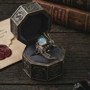 May include: A bronze-toned dragon ring with a large, opalescent orb held in its claws, displayed in an octagonal, velvet-lined box. The box is decorated with engraved symbols and a dragon motif, suggesting a fantasy or gothic theme.