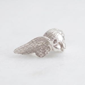 May include: A silver-colored angel wing and head sculpture. The sculpture features detailed feathering on the wing and a serene facial expression. The angel's head is turned slightly to the right, with the wing extending to the left.