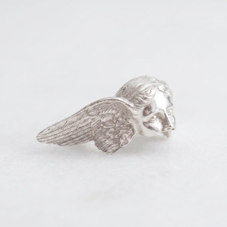 May include: A silver-colored angel wing and head sculpture. The sculpture features detailed feathering on the wing and a serene facial expression. The angel's head is turned slightly to the right, with the wing extending to the left.