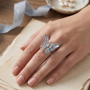 May include: A silver ring featuring a light blue butterfly design with a clear gemstone center. The butterfly's wings have a delicate, openwork pattern. The ring is shown on a hand with well-manicured nails.