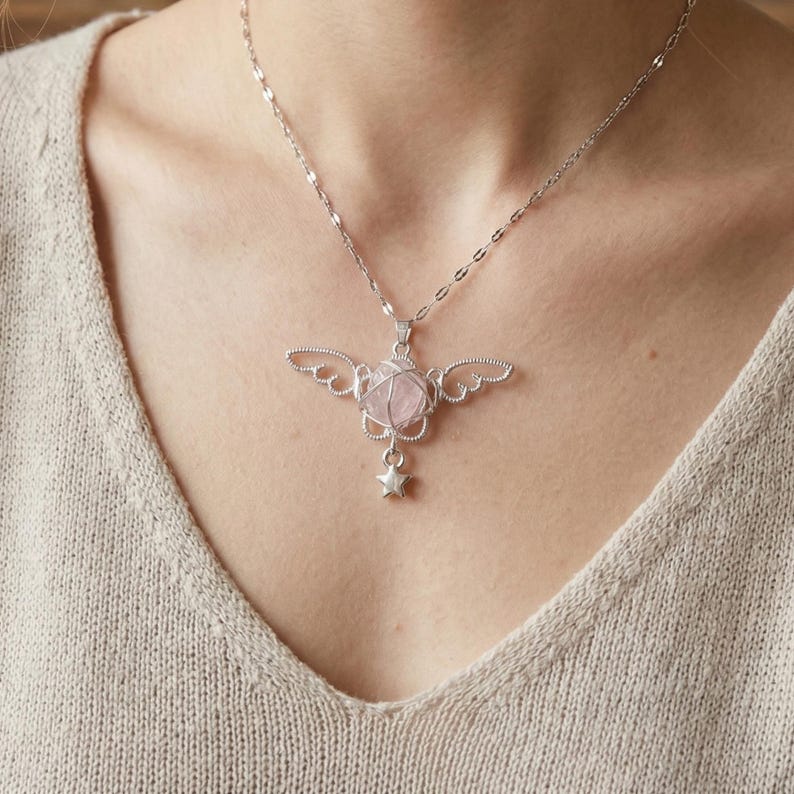 May include: A silver-toned necklace featuring a pendant with a pink gemstone encased in wire, adorned with wing-shaped details and a dangling star charm. The necklace is worn on a person.