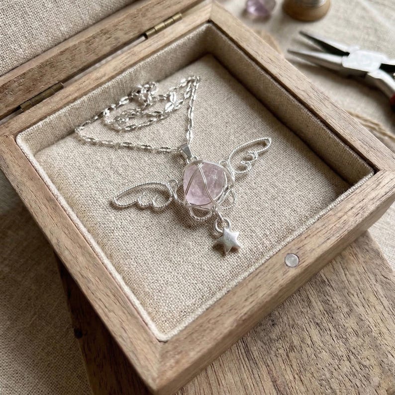 May include: A silver necklace with a pink gemstone pendant, shaped like a heart and wire-wrapped, is displayed in a wooden box. The pendant has silver wings and a star charm. The box is lined with beige fabric.