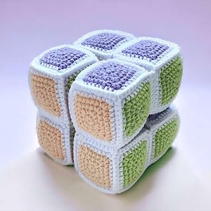 May include: A handmade crochet cube puzzle in a variety of colors. The cube is made of individual crochet squares in shades of lavender, green, and tan, with white borders. The cube is a unique and colorful handmade toy.
