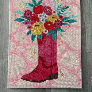 Puede incluir: Una obra de arte decorativa que presenta una bota de vaquero rosa llena de un vibrante ramo de flores. Las flores incluyen flores rojas, amarillas y rosas con hojas verdes. El fondo es un patrón de puntos rosas y blancos.