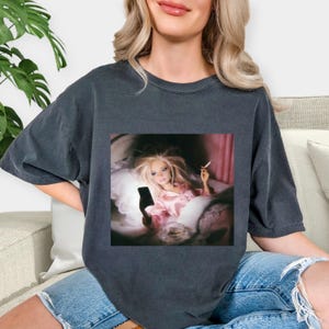 May include: Dark gray t-shirt featuring a square image of a doll in bed, holding a phone and a cigarette. The doll is wearing a pink satin pajama set. The t-shirt is a relaxed fit.