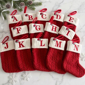 Könnte beinhalten: Eine Sammlung roter Weihnachtsstrümpfe aus Zopfstrick mit weißen Bündchen. Jeder Strumpf ist mit einem roten gestickten Buchstaben von A bis N und einer kleinen gestickten Weihnachtsmütze darüber versehen. Jeder Strumpf hat eine rote Aufhängeschlaufe.
