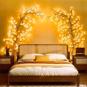 Puede incluir: Dormitorio con cama de madera y cabecero tejido. La cama está hecha con ropa de cama blanca y amarilla. Dos mesitas de noche flanquean la cama. Luces de cadena en forma de ramas adornan la pared sobre la cama, creando un ambiente cálido.
