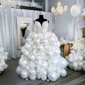 May include: A white balloon dress on a black mannequin. The bodice is covered in small white beads, and the skirt is made of various sizes of white balloons. The dress is displayed in a room with white curtains, a mirror, and chandeliers.