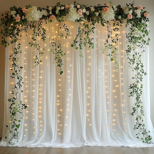 May include: A white fabric backdrop curtain adorned with a top garland of white hydrangeas, peach roses, and green ivy vines. Cascading ivy branches hang down the sides, interspersed with vertical strings of warm white fairy lights.