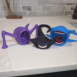 May include: Three articulated 3D printed figures: a purple creature with long limbs and a hollow body, a black hammerhead-shaped creature with multiple blue eyes and sharp white teeth, and a blue shark-like creature with an open mouth and red interior.