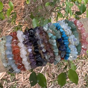 May include: A collection of gemstone chip bracelets in various colors, including green, blue, purple, and red. The bracelets are displayed on a tan fabric roll, with green foliage in the background. Each bracelet is made of small, irregular-shaped stones.