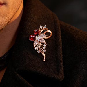 Puede incluir: Broche floral dorado con detalles de piedras preciosas rojas y cristales blancos brillantes. El broche presenta un diseño floral con un lazo y delicados detalles de hojas. Un accesorio elegante.