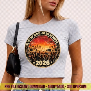 May include: A pale grey crop top with a circular graphic. The graphic includes the text "WE GOT THE PEOPLE WE GOT THE POWER" and a crowd silhouette with a cityscape. The year "2026" is below. The shirt is worn by a person.