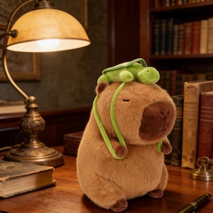 May include: A plush capybara toy in shades of brown with a small green turtle on its head. The capybara has a soft, furry texture and a friendly expression. A green strap is attached to the turtle and drapes down the capybara's sides.