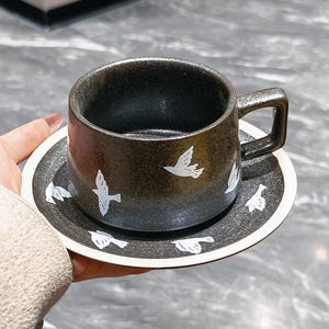 Pode incluir: Um conjunto de três canecas de cerâmica com uma cobertura salpicada de cor creme. Cada caneca apresenta um design pintado à mão diferente: uma folha azul e verde, uma flor amarela e uma baga vermelha. As canecas têm um design simples e moderno.