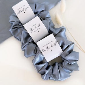 May include: Three dusty blue satin hair scrunchies, each wrapped in a white paper band. The bands feature black text: I CAN'T SAY I DO without you, I CAN'T TIE the knot WITHOUT YOU, and WILL YOU HELP ME tie the knot? Each includes a small black heart.