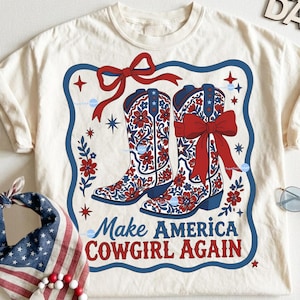 Puede incluir: Camiseta color crema con un dise&ntilde;o de dos botas de vaquero con patrones florales rojos y lazos. Las botas est&aacute;n sobre un borde azul con el texto "Make America Cowgirl Again" en rojo y azul.