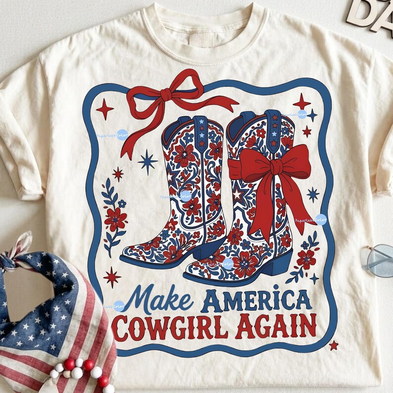 Puede incluir: Camiseta color crema con un dise&ntilde;o de dos botas de vaquero con patrones florales rojos y lazos. Las botas est&aacute;n sobre un borde azul con el texto "Make America Cowgirl Again" en rojo y azul.