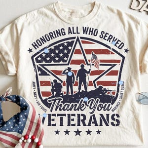 Puede incluir: Camiseta de color crema con un dise&ntilde;o patri&oacute;tico. El gr&aacute;fico presenta un motivo de estrellas y rayas con siluetas de soldados y el texto "Honoring All Who Served" y "Thank You Veterans".