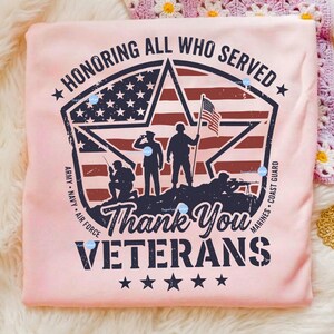 Puede incluir: Una camiseta rosa claro con un dise&ntilde;o patri&oacute;tico. El dise&ntilde;o incluye un emblema en forma de estrella con la bandera estadounidense, siluetas de soldados y el texto "HONORING ALL WHO SERVED" y "Thank You VETERANS".