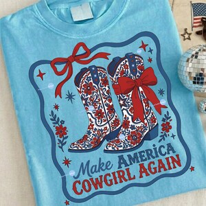 Puede incluir: Camiseta azul claro con un dise&ntilde;o de dos botas de vaquero con lazos rojos y estampados florales. Las botas est&aacute;n enmarcadas en azul y rojo, con el texto "Make America Cowgirl Again" debajo.