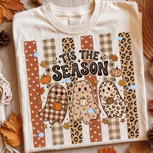 May include: A cream-coloured t-shirt featuring a graphic with the text 'TIS THE SEASON' in dark brown letters. The design includes three ghosts with different patterns, including plaid, solid, and leopard print, each holding a small pumpkin.