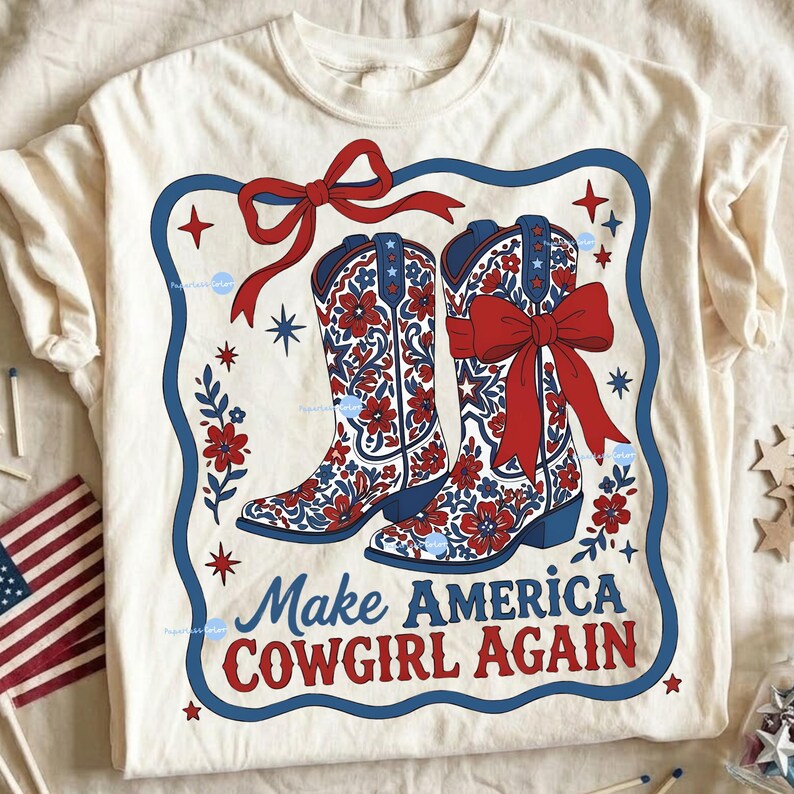 Puede incluir: Camiseta color crema con un dise&ntilde;o de dos botas de vaquero con patrones florales rojos, detalles azules y lazos rojos. La frase "Make America Cowgirl Again" est&aacute; impresa en rojo y azul. El dise&ntilde;o est&aacute; enmarcado con un borde azul y estrellas.