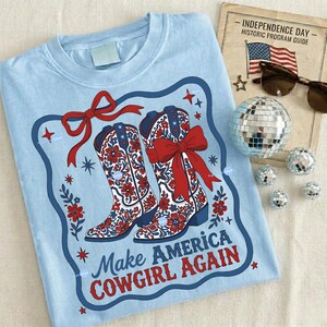 Puede incluir: Camiseta azul claro con un gr&aacute;fico de botas de vaquero florales rojas y blancas con lazos rojos, y el texto "Make America Cowgirl Again". El dise&ntilde;o est&aacute; enmarcado en un borde azul con estrellas y detalles florales.