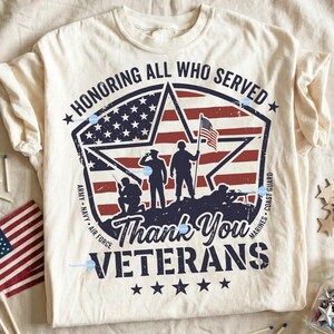 Puede incluir: Camiseta de color crema con un dise&ntilde;o patri&oacute;tico. El gr&aacute;fico incluye un emblema en forma de estrella con la bandera estadounidense, siluetas de soldados y el texto "Honoring All Who Served" y "Thank You Veterans".