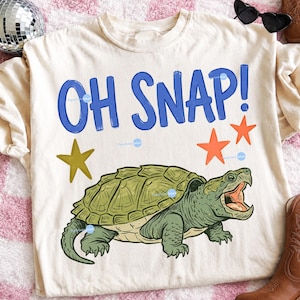 Könnte beinhalten: Cremefarbenes T-Shirt mit einer Comic-Schnappschildkröte mit offenem Maul. Der blaue Text "OH SNAP!" befindet sich darüber, mit orangefarbenen und gelben Sternen. Das Shirt hat lange Ärmel.