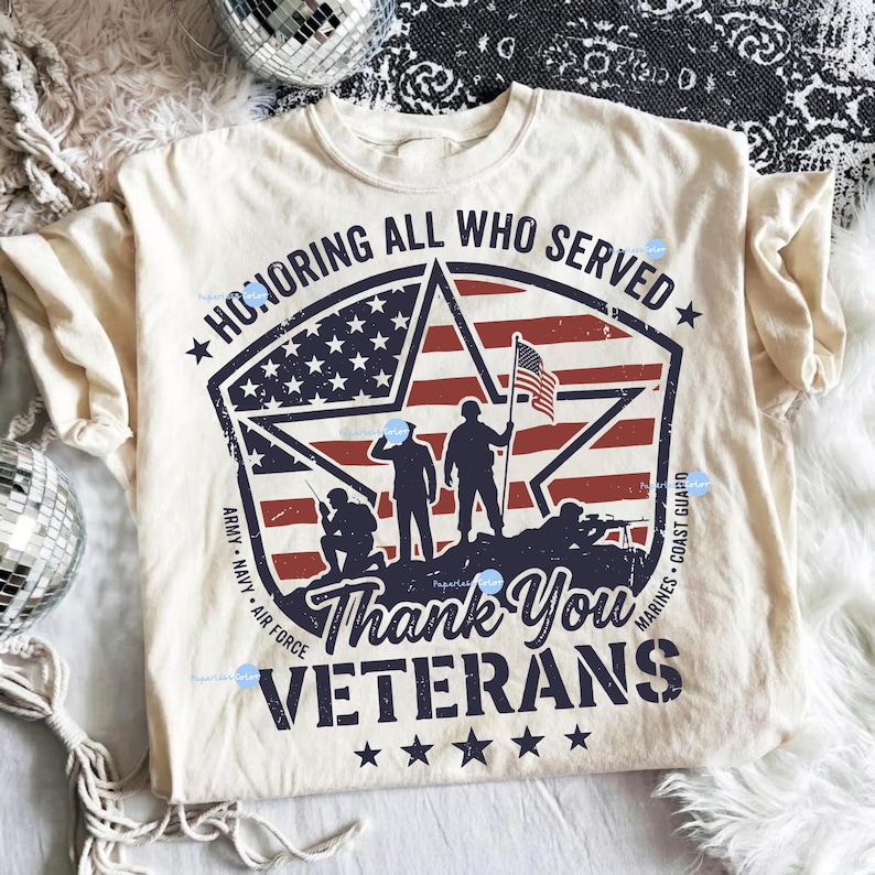 Puede incluir: Camiseta color crema con un dise&ntilde;o patri&oacute;tico. El gr&aacute;fico presenta una estrella con la bandera estadounidense, siluetas de soldados y el texto "Honoring All Who Served" y "Thank You Veterans".