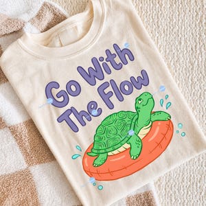 Puede incluir: Sudadera color crema con la frase "Go With The Flow" en letras moradas. Una tortuga de dibujos animados, con caparazón verde, flota en un flotador naranja. El diseño incluye gotas de agua azules.