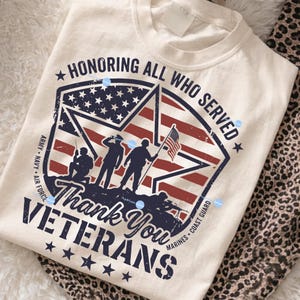 Puede incluir: Camiseta color crema con un dise&ntilde;o patri&oacute;tico. El gr&aacute;fico presenta una bandera estadounidense en forma de estrella con siluetas de soldados y el texto "Honoring All Who Served" y "Thank You Veterans."