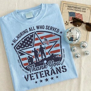 Puede incluir: Camiseta azul claro con un dise&ntilde;o patri&oacute;tico. El gr&aacute;fico presenta una estrella y rayas en rojo, blanco y azul, con siluetas de soldados y el texto "Thank You Veterans". Tambi&eacute;n se muestran gafas de sol, una bola de discoteca y un programa vintage del D&iacute;a de la Independencia.