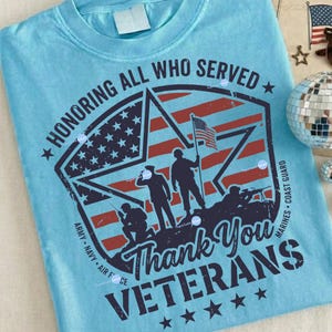 Puede incluir: Una camiseta azul claro con un dise&ntilde;o patri&oacute;tico. El gr&aacute;fico presenta un emblema en forma de estrella con la bandera estadounidense, siluetas de soldados y el texto "Honoring All Who Served" y "Thank You Veterans."