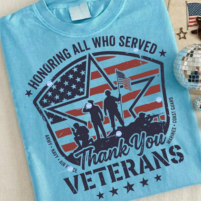 Puede incluir: Una camiseta azul claro con un dise&ntilde;o patri&oacute;tico. El gr&aacute;fico presenta un emblema en forma de estrella con la bandera estadounidense, siluetas de soldados y el texto "Honoring All Who Served" y "Thank You Veterans."