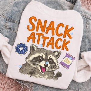 Op de afbeelding: Wit sweatshirt met de woorden "SNACK ATTACK" in oranje blokletters. Een cartoon wasbeer met een roze tong en een blauwe donut-afbeelding zijn ook te zien. Een zak chips is ook afgebeeld.