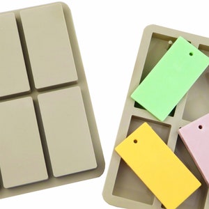 Op de afbeelding: Twee siliconen mallen voor het maken van rechthoekige harsambachten. De mallen hebben een gat voor een sprongring. Eén mal is lichtbruin en de andere is grijs. De mallen zijn gevuld met kleurrijke harsstukken.