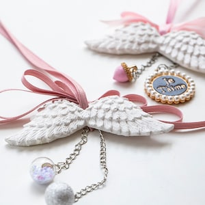Può includere: Due ali d'angelo bianche con un nastro rosa e un ciondolo con le parole "Je T'aime" in una cornice incrostata di perle.